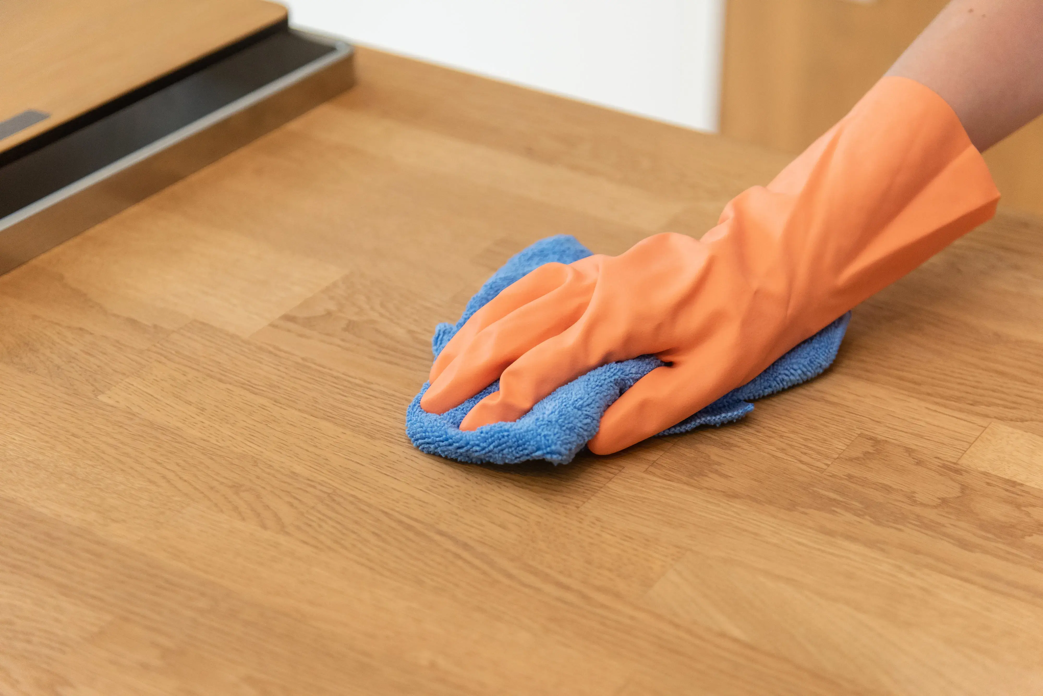 Hand mit orangem Handschuh wischt Holztisch mit blauem Tuch ab.